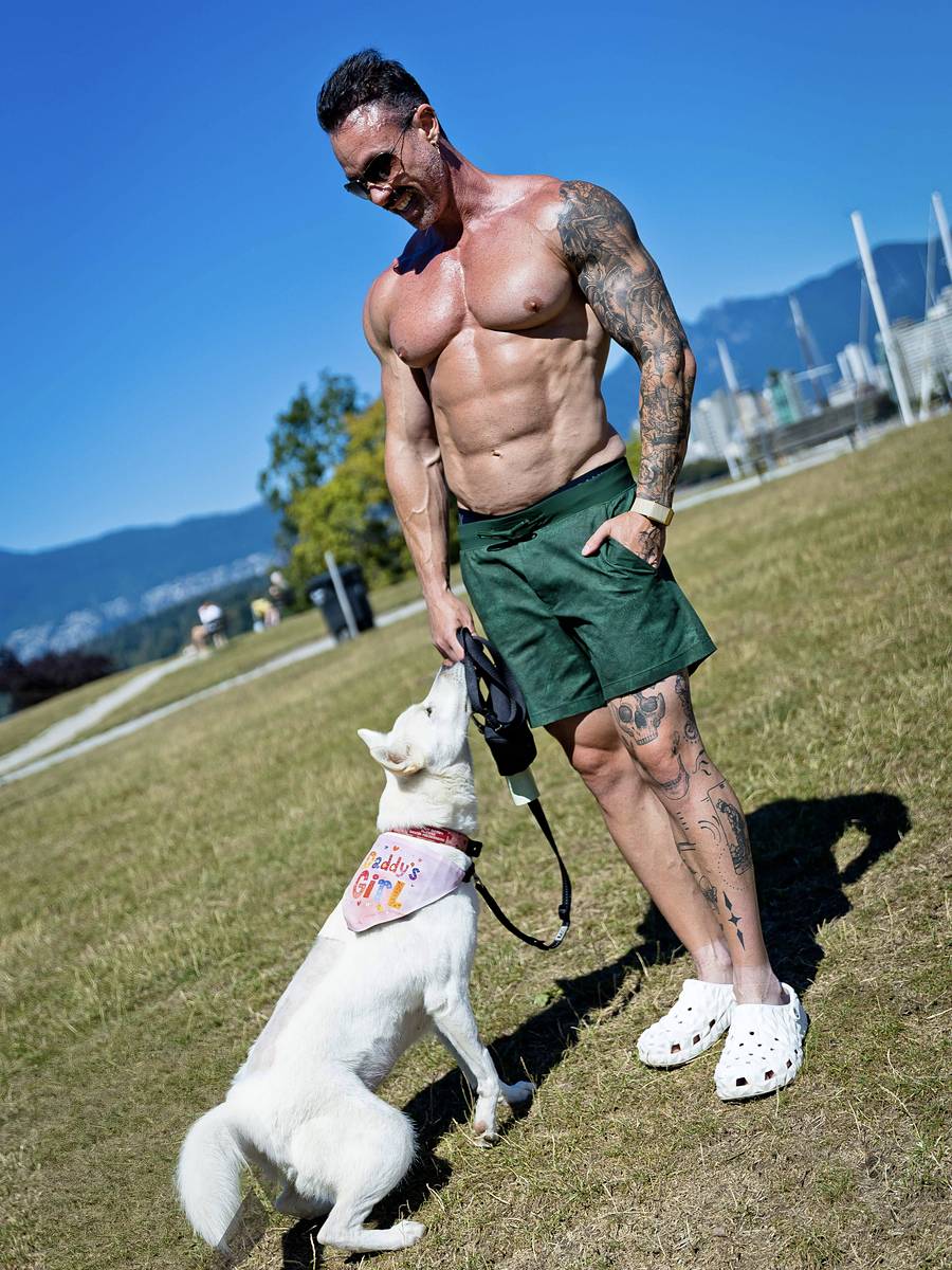 David Turnbull standing on a grassy hill in green shorts and crocs while his white dog Juhee in a pink bandana reaches up to him