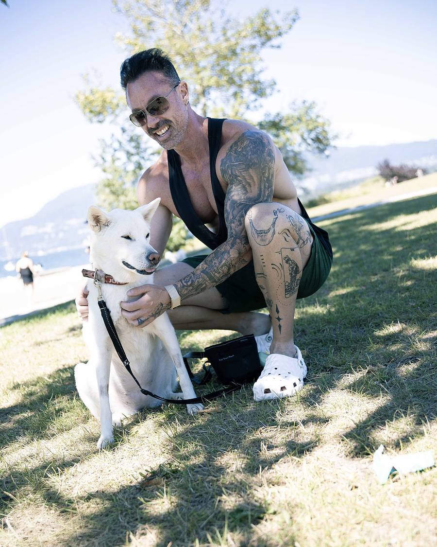 David Turnbull in a tank top and sunglasses crouching on the grass with his white dog at a Vancouver beach park